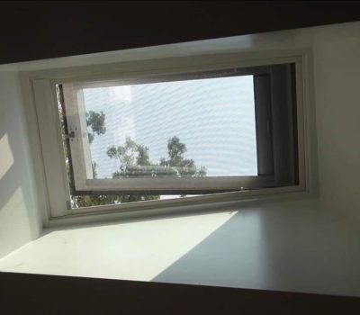 A skylight window with a mesh screen partially open, showing a blue sky and tree branches outside, casting shadows on the walls.