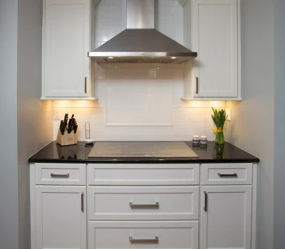 Modern kitchen with white cabinets, stainless steel range hood, black countertop, and induction cooktop. Knife block and flower vase for decor.
