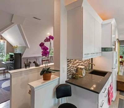 A modern loft-style interior with a desk, plants, and orchids. Bright space features a kitchen area and slanted ceilings with skylights.