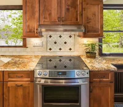 A modern kitchen features wooden cabinets, a stainless steel oven, granite countertops, tile backsplash, and sunlight through large windows with greenery outside.