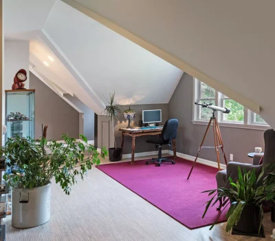 A cozy attic office with a desk, computer, telescope, plants, and decorative lamp, highlighted by a purple rug under sloped ceilings.