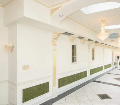 Elegant interior hallway with ornate columns, gold accents, and chandeliers. White walls are complemented by green wall textures, creating a luxurious atmosphere.