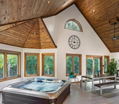 Spacious room with wooden ceiling, hot tub, large windows, wall clock, table with benches, and lush greenery outside. Cozy, inviting atmosphere.