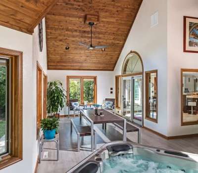 Inviting sunroom with a wooden ceiling, bubbling hot tub, seating area, plants, and large windows offering a view of lush greenery.
