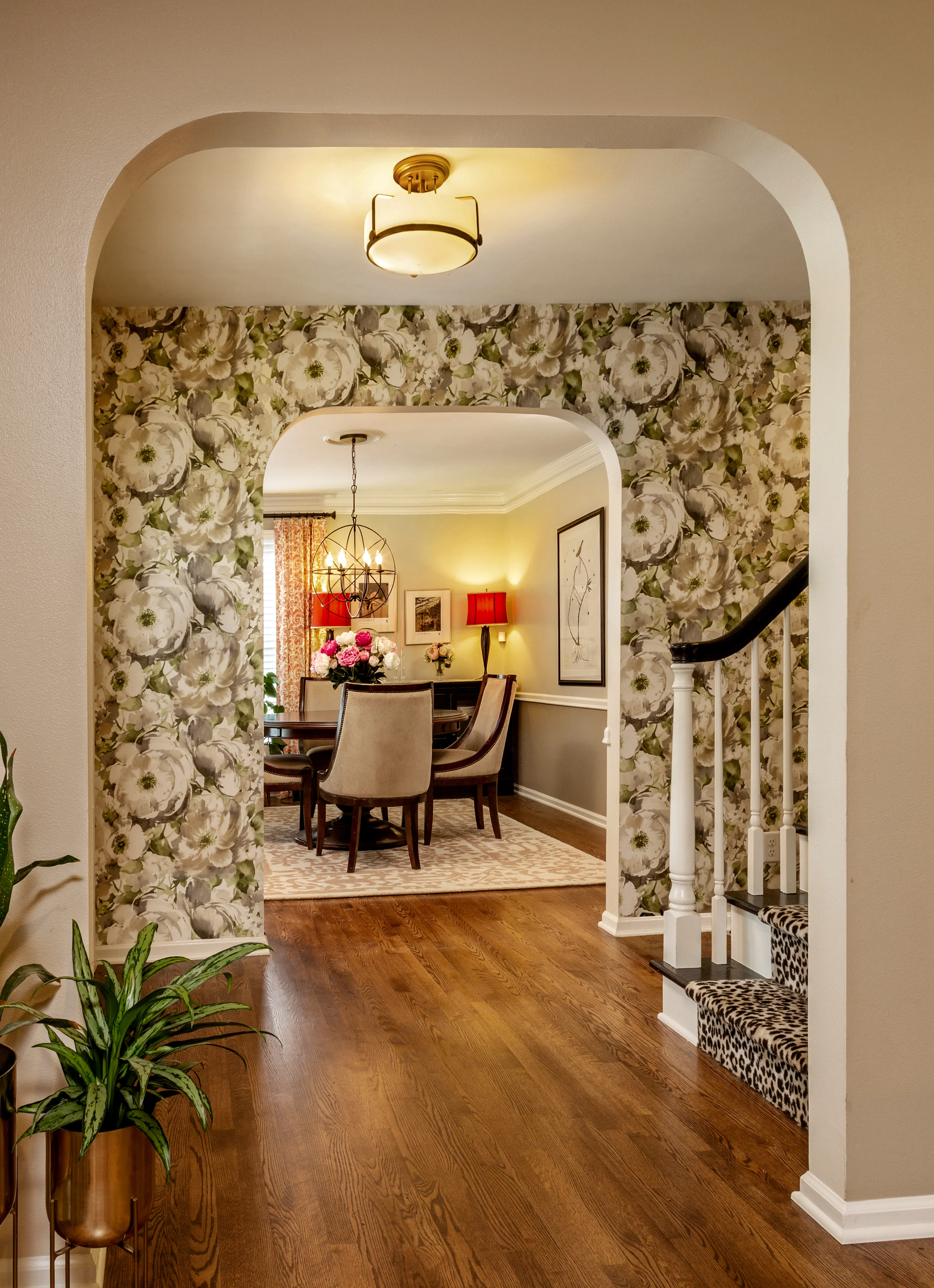 Arched hallway leads to a cozy dining room with floral wallpaper, elegant chandelier, patterned chairs, and decorative table setting. Hardwood floors and artwork complete.