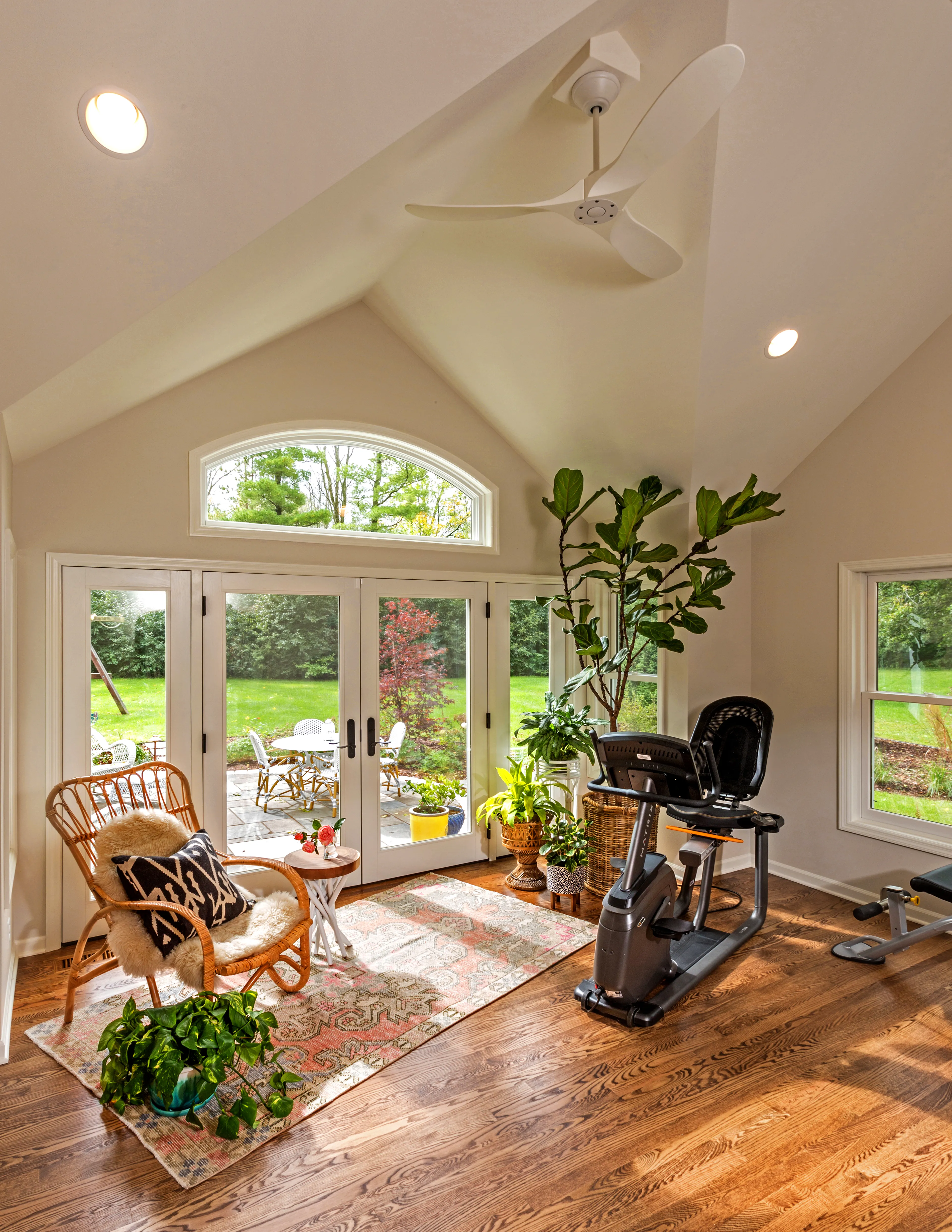 A sunlit room features a cozy seating area, exercise bike, wooden floor, vibrant plants, and large windows opening to a lush green garden.