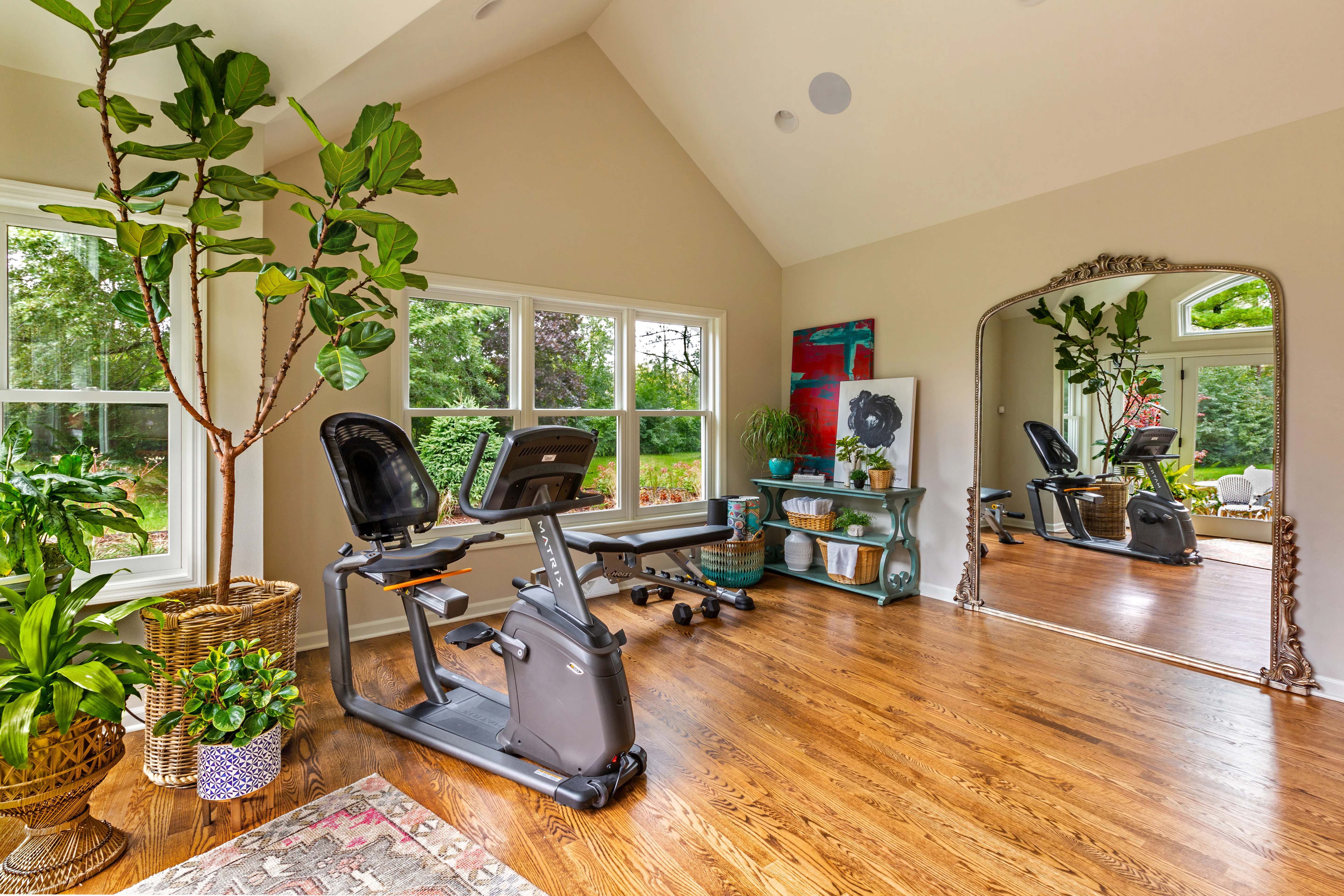 Bright home gym with exercise equipment, large mirror, plants, and colorful artwork. Hardwood floors and window view create an inviting atmosphere.