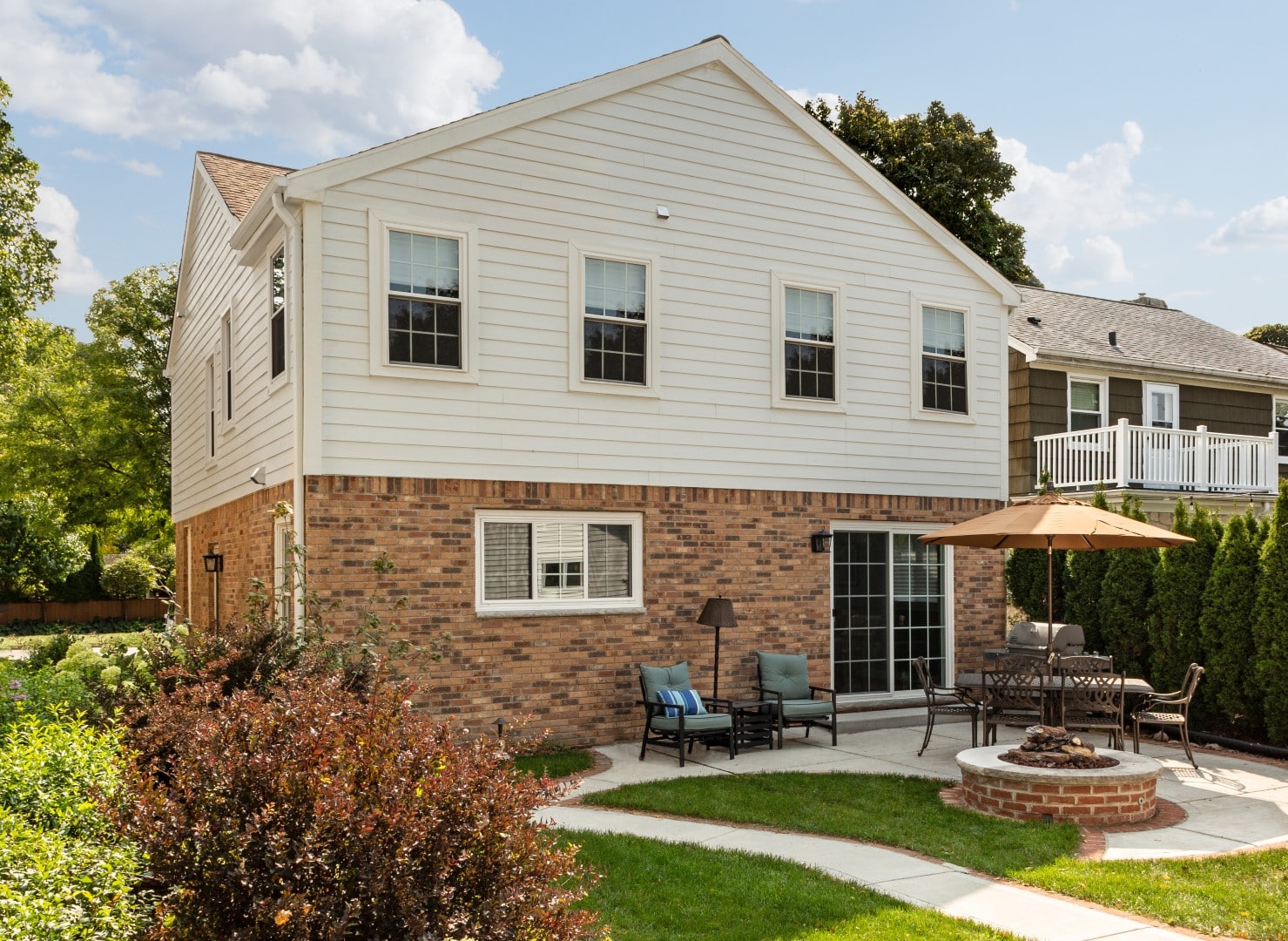 A two-story house with a brick and siding exterior, patio furniture, umbrella, and fire pit in a landscaped backyard setting.