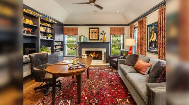 Cozy living room with a fireplace, bookshelves, and a patterned red rug. Art decorates the walls, complementing the warm, inviting atmosphere.