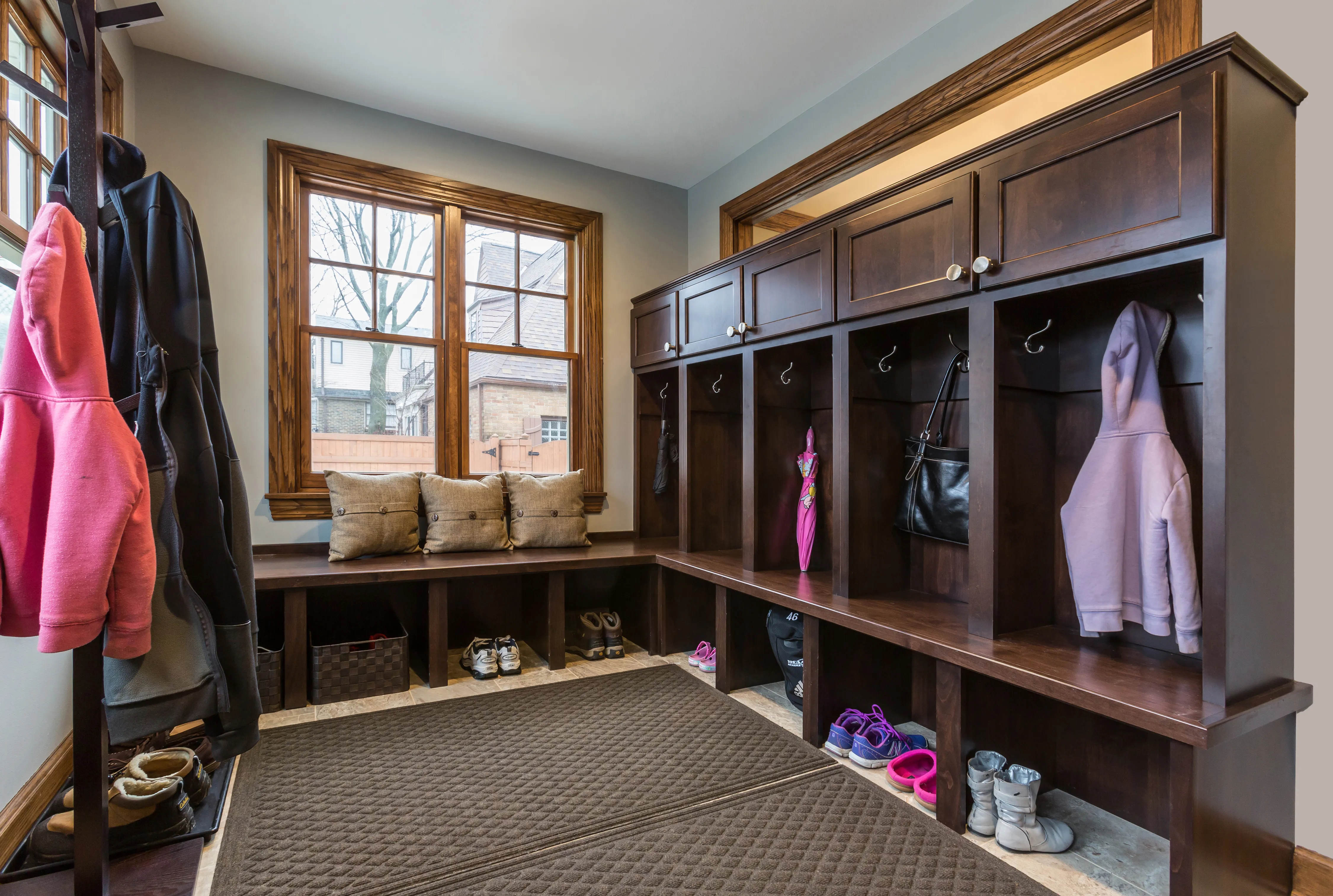 A tidy mudroom with wooden cubbies, coats, shoes, a pink umbrella, and pillows. Large windows overlook an outdoor scene with trees.