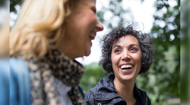Two people are smiling and talking outdoors, surrounded by green trees. The atmosphere is joyful and relaxed, with natural lighting.