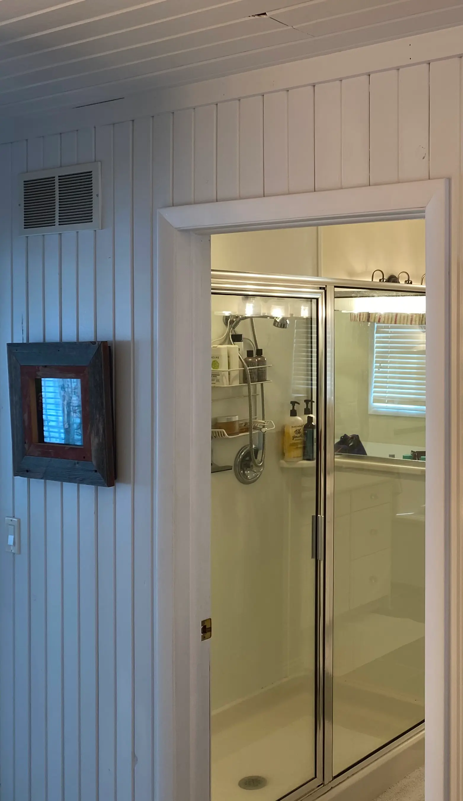 A glass shower door in a modern bathroom, with shelves holding toiletries, next to a framed mirror on wooden walls.