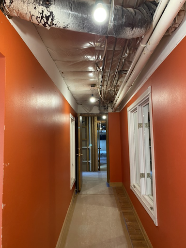Narrow hallway with vibrant orange walls and exposed ceiling pipes, leading to an open door. Bright lighting enhances the industrial aesthetic.