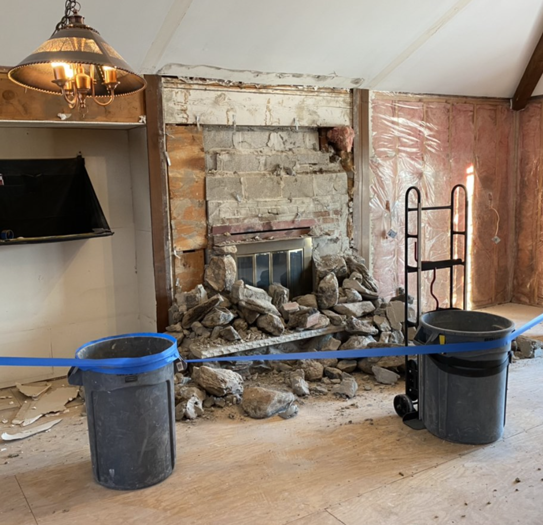 Room under renovation with exposed insulation, partially demolished fireplace, broken rock debris, plastic tarp, blue tape, two bins, dolly, and a chandelier.